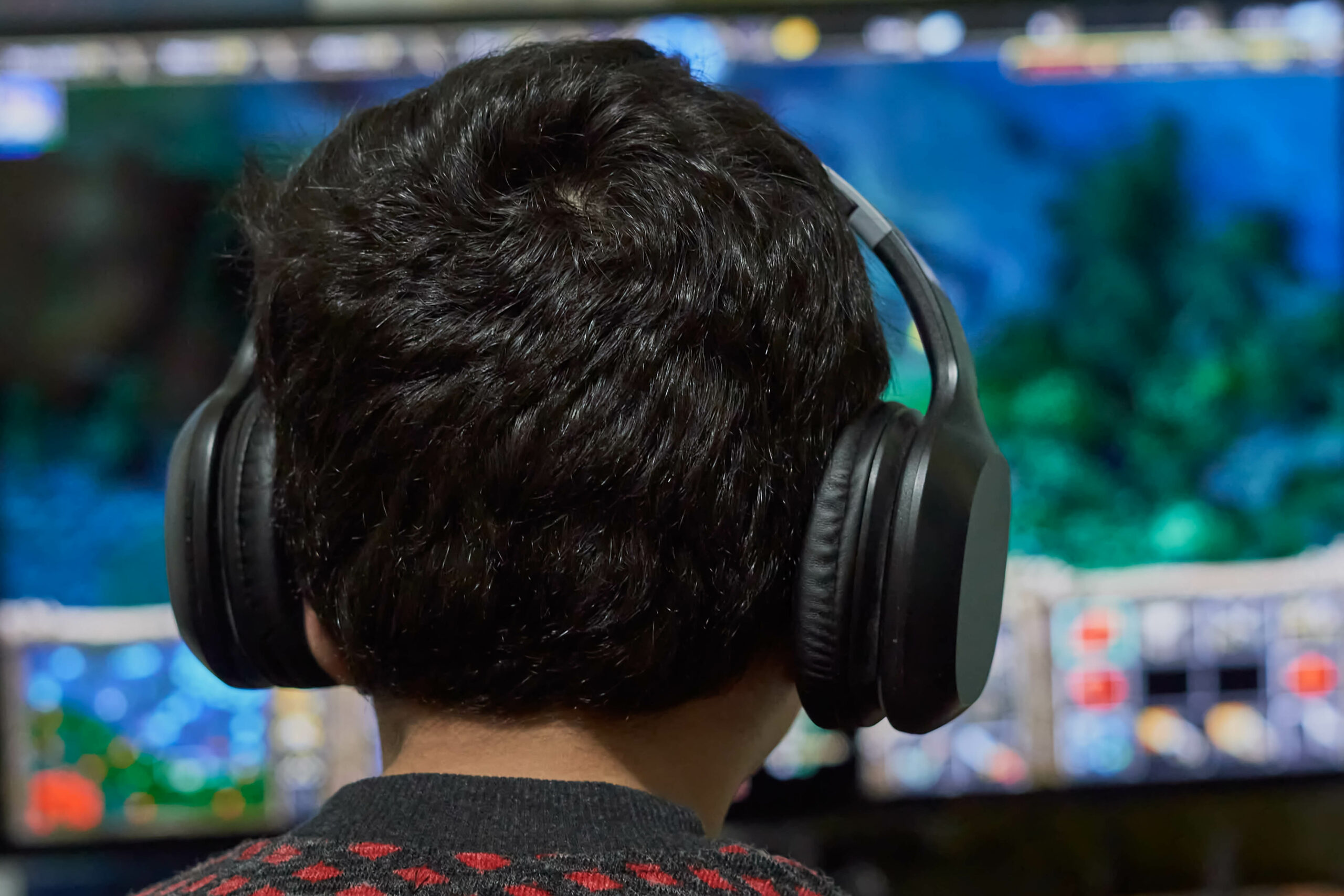 Foto: Im Vordergrund ist der Hinterkopf eines Kindes mit Kopfhörern. Es spielt ein Computerspiel, dessen Inhalt unscharf im Hintergrund auf einem Bildschirm zu sehen ist.