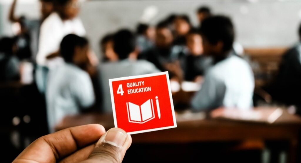 Ein Symbolbild für BNE: Eine Hand hält ein Kärtchen mit dem SDG "Qualiy Education" in die Höhe. Im Hintergrund ist eine Gruppe Lernender zu sehen.